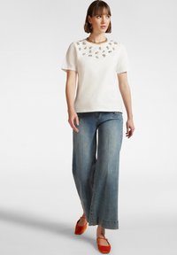 T-shirt bianca in cotone con decorazioni in argento attorno al collo, abbinata a jeans in denim a gamba larga blu chiaro e ballerine arancioni.