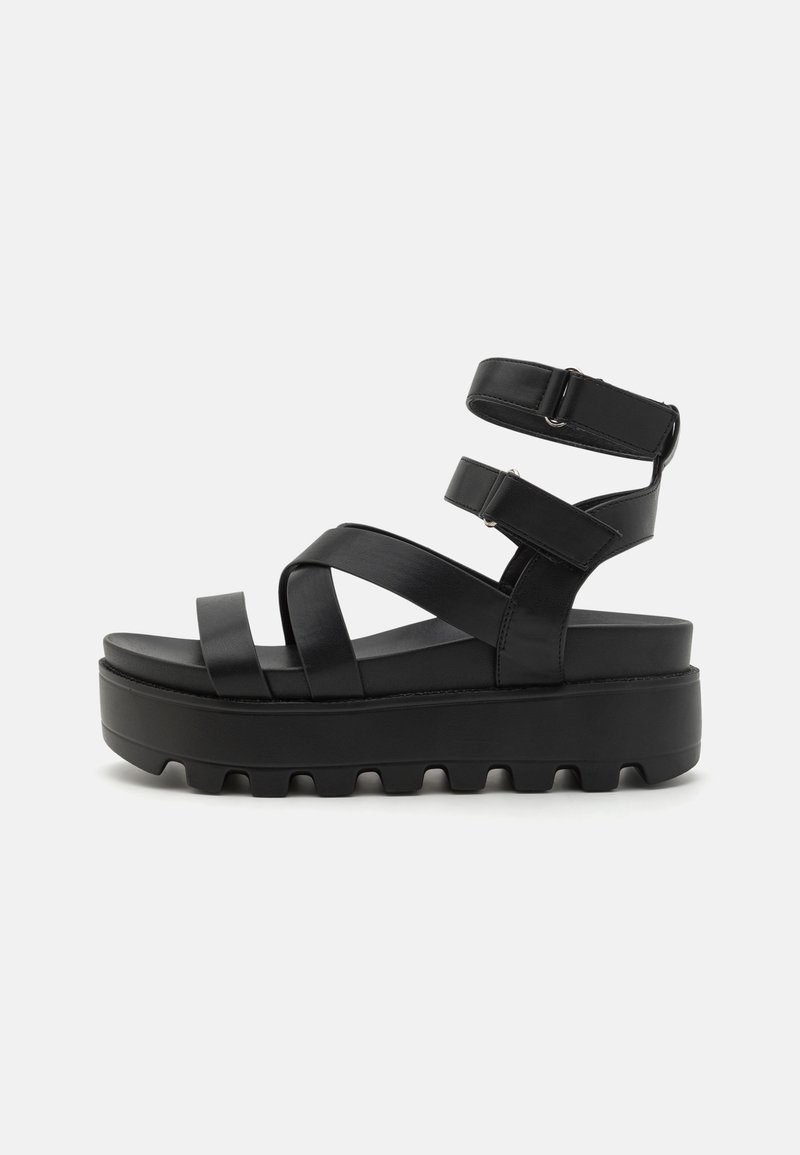 Black platform sandals featuring cross-over straps, adjustable ankle buckles, and a textured sole for grip. Made from synthetic material.