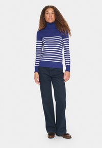 Pull à col roulé bleu marine avec des rayures blanches horizontales, ajusté au niveau du buste et ample en bas, associé à un pantalon large sombre.