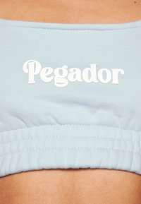 Jasnoniebieski krótki top z prążkowanej tekstury, z białym haftowanym napisem "Pegador". Posiada zgromadzony elastyczny dół dla dopasowanego wyglądu.