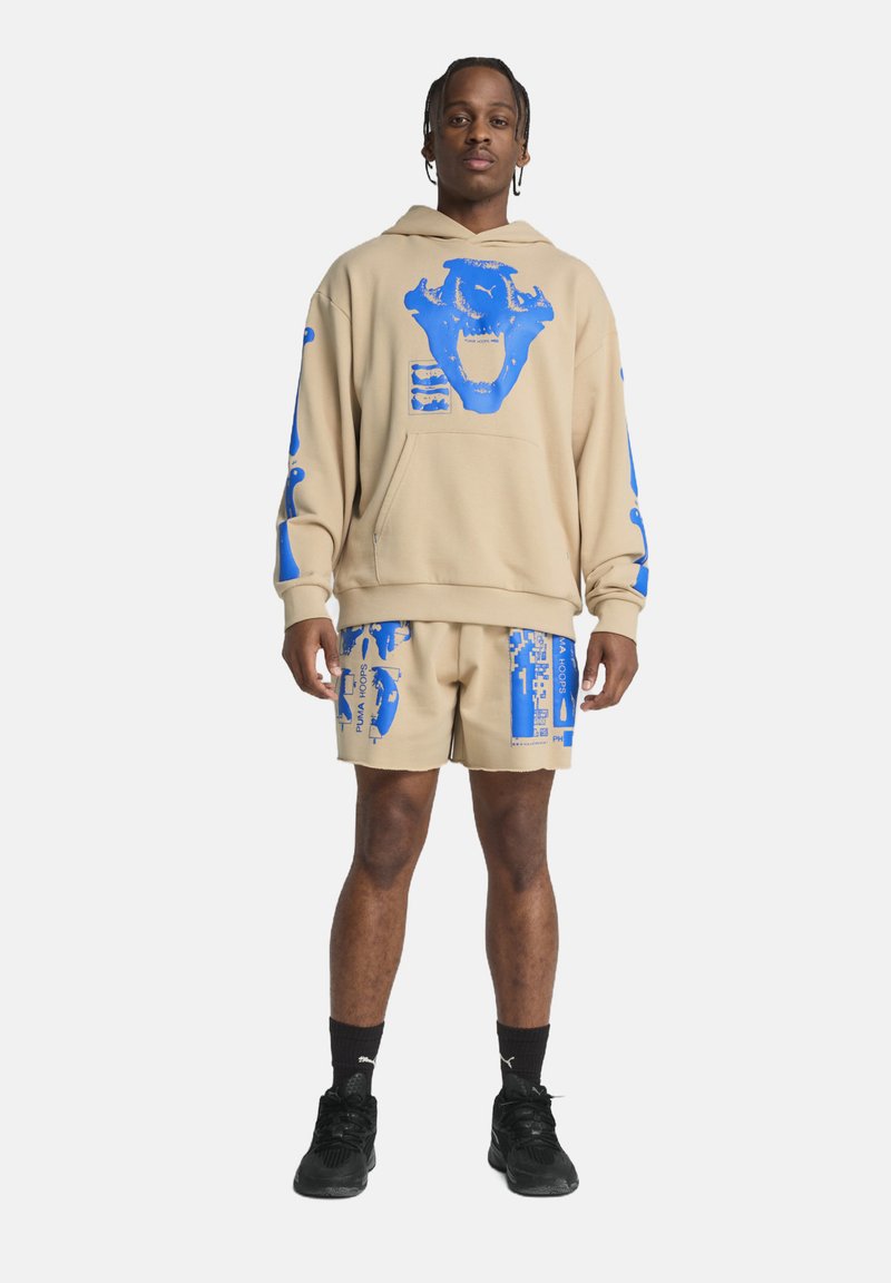 Sudadera con capucha beige con diseños gráficos azules y pantalones cortos a juego, con patrones abstractos y texto impreso. Combinado con zapatillas negras.