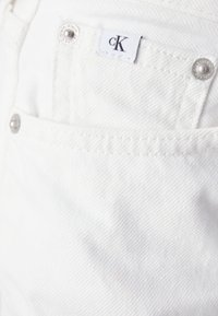 Λευκά τζιν παντελόνια διαθέτουν ορατό μπροστινό τσέπη με ετικέτα "cK", ασημένια κουμπιά και υφή υφάσματος.