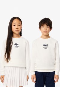 Hvit bomullsgenser med ribbestrikkede mansjetter, med en mørkeblå Lacoste-logo. Jenta har på seg et hvitt plissert skjørt; gutten har på seg mørke bukser.