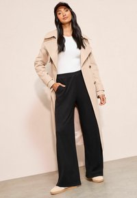 Beige tranšejkots pāri baltai augšai, kombinēts ar melniem platām biksēm un beigu kurpēm. Gluda auduma, strukturēts dizains un priekšējie kabatas.