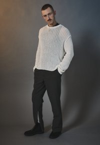 Suéter grueso de punto blanco con textura acanalada, combinado con pantalones oscuros a medida que tienen cremalleras laterales y calzado negro.
