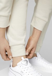 Pantalones sudados de color crema con puños acanalados, combinados con zapatillas blancas que presentan un logotipo metálico y acentos texturizados en la lengüeta.
