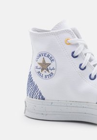 Sapatilha alta de lona branca com logotipo bordado azul e dourado, ilhós em contraste e detalhamento em costura texturizada no calcanhar.