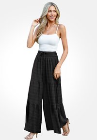 Zwarte, wijde broek met een textuurpatroon en een elastische tailleband, gecombineerd met een witte tanktop en beige sandalen.