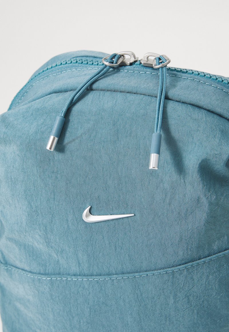 Mochila de color azul claro con tejido texturizado, cierre de cremallera doble y logotipo plateado de Nike. Presenta cordones de ajuste y un acabado suave.