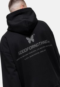 Hombre con sudadera negra con capucha que tiene un logo de mariposa y el texto "GOODFORNOTHING" en la parte de atrás, junto con una frase sobre la valentía y Manchester, Inglaterra.