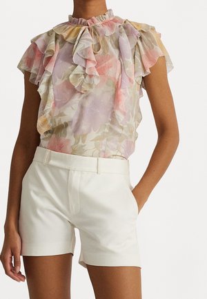 Kvinde iført en blomsterprintet transparent bluse med flæser og hvide højtaljede skræddersyede shorts, den ene hånd i lommen, mod en hvid baggrund.