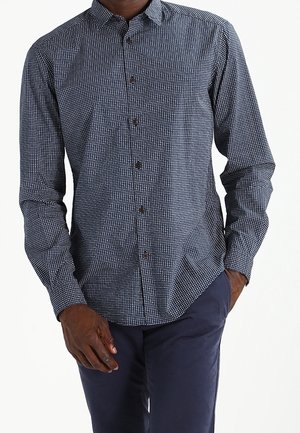 Homme portant une chemise à manches longues à carreaux bleu marine et blanc boutonnée, avec un pantalon bleu foncé, une main dans la poche, fond blanc uni.