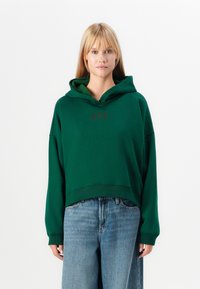 Haut à capuche cropped vert en tissu doux avec une grande capuche. Présente un logo "GAP" discret en impression foncée et des manches amples. Porté avec un pantalon en denim.