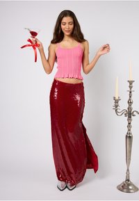 Różowy prążkowany krótki top z falistymi brzegami, w połączeniu z długą, cekinową czerwoną spódnicą. Modelka trzyma koktajl z wstążką. Wyeksponowane srebrne szpilki.