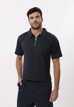Uomo che indossa una polo blu navy a maniche corte con cerniera frontale e pantaloni blu navy abbinati, in piedi davanti a uno sfondo bianco.