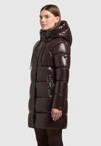 Kvinde iført en lang, mørkebrun skinnende pufferjakke med hætte og sorte snore, stående sidelæns mod en ensfarvet baggrund.