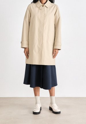 Personne portant un manteau beige jusqu'aux genoux sur une jupe bleu marine, des chaussettes crème et des mocassins à plateforme blancs sur un fond clair uni.