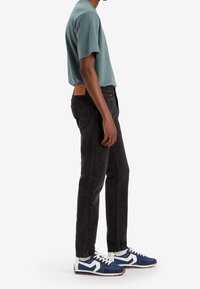 Jeans de mezclilla negra, camisa de manga corta teal y zapatillas azules con acentos blancos. Presenta una etiqueta en la cintura y el dobladillo vuelto.