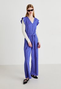 Paarse jumpsuit met zigzagpatroon, ceintuur in de taille, rimpels in de mouwen en knopen. Ggedragen over een witte, gestructureerde blouse met lange mouwen en zwarte schoenen.