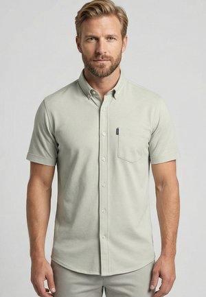 Blonde man met baard die een lichtgrijs overhemd met korte mouwen en borstzak draagt, staand tegen een effen achtergrond.