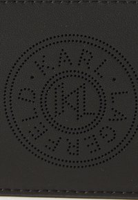 Accessorio in pelle nera con logo circolare perforato che riporta "KARL LAGERFELD" e un design di raggi di sole, texture liscia e bordi cuciti.
