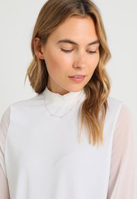 Weiße Bluse mit rüschenbesetztem hohen Kragen, durchsichtigen langen Ärmeln und glatter Textur. Kombiniert mit einer zarten silbernen Halskette, die kleine Perlen enthält.