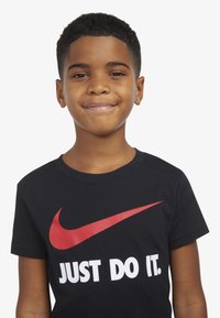 Melnas kokvilnas T-krekls ar lielu sarkano Nike zvīņu un baltu tekstu "TIKAI DARI TO." Īsas piedurknes un apaļas kakla dizains.
