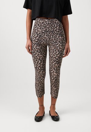 Femme portant un pantalon court taille haute à imprimé léopard, assorti à des ballerines noires et un top court noir, posant devant un fond uni.