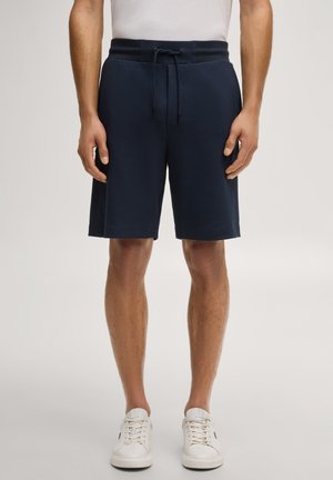 Mężczyzna w granatowych szortach z troczkiem i białych tenisówkach, stojący na jednolitym jasnym tle.