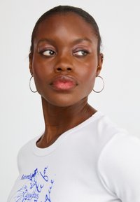 Hvid bomulds T-shirt med rund hals og blå grafisk tryk. Modellen har kort mørkt hår, bærer hooped øreringe og har en neutral makeup. 