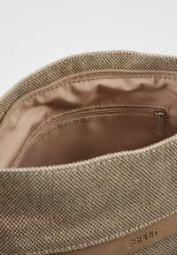 Tygkasse i beige med strukturerad väv. Insidan har en slät, ljusbrun foder och en liten ficka med dragkedja. Logotyp synlig.