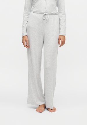 Persoon die lichtgrijze wijde broek met koordje in de taille en een shirt met lange mouwen draagt, staat op blote voeten op een witte achtergrond.