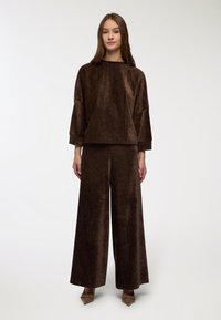 Set coordinato in velluto marrone composto da un top squadrato con maniche larghe e collo alto, abbinato a pantaloni a gamba larga. Texture liscia e vestibilità comoda.