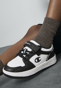 Baskets noir et blanc avec une tige en cuir texturé, bout rond et bande Velcro. Logo "Champion" présent sur le côté et la bande.