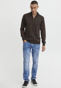 Pullover lavorato a maglia in marrone scuro con sfumature arancioni, dotato di colletto con mezza zip e orlo a coste. Indossato con jeans azzurri chiari e sneakers nere.