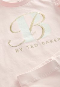 Rozā kokvilnas krekls ar garām piedurknēm, kurā ir zeltīts izšuvums ar "B" logotipu un tekstu "BY TED BAKER", ar gludu tekstūru.