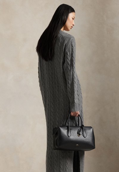 Kvinde i en lang grå kabelstrikket sweaterkjole, der holder en sort lædertaske mod en ensfarvet beige baggrund.