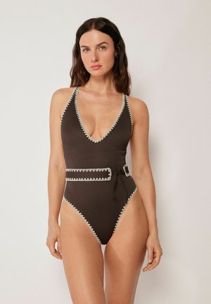 Frau mit langen dunklen Haaren, die einen ärmellosen dunkelbraunen texturierten Badeanzug mit weißem Besatz und passendem Gürtel trägt, steht vor einem schlichten Hintergrund.