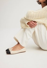 Ballerines beiges avec un embout noir et un accent en nœud, présentant une finition lisse semblable au cuir et une forme simple et arrondie.