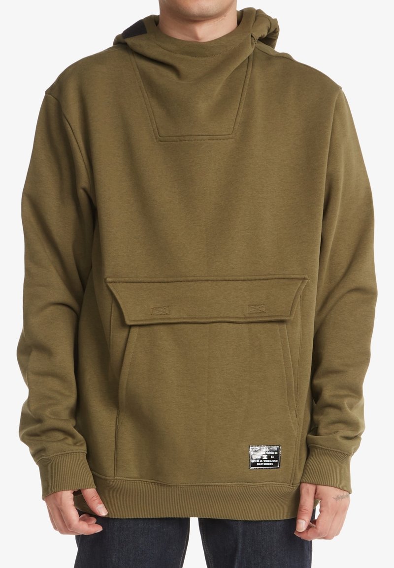 Homme portant un sweat à capuche vert olive avec une poche frontale et un petit patch noir et blanc près de l'ourlet.