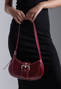 Frau in einem schwarzen Kleid, die eine kleine burgunderfarbene Schultertasche aus Leder mit einer Schnallendetail und Reißverschluss hält.