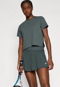 Sötétzöld sportos szett, amely egy testhezálló, rövid ujjú pólóból és egy passzoló, bővülő szoknyából áll, könnyű anyagból készül.