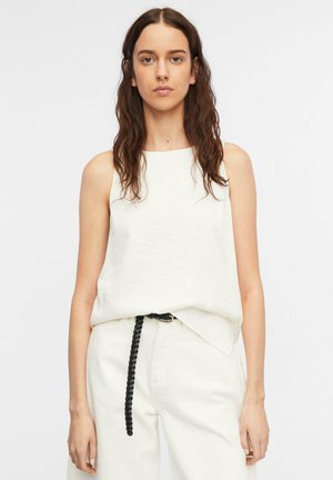 Kobieta o długich ciemnych włosach, ubrana w biały top bez rękawów, białe spodnie z wysokim stanem i czarny pleciony pasek na jednolitym tle.