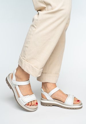 Person wearing beige corduroy pants and white sandals with stud detailing, showing painted red toenails on white background.