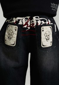 Rückansicht von schwarzen Denim-Jeans mit weißen Ace of Spades Totenkopf-Patches auf beiden Gesäßtaschen und einem schwarzen Top mit weißem und rotem Design.