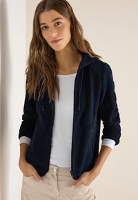 Jeune femme aux longs cheveux portant un sweat à capuche bleu marine zippé sur un t-shirt blanc et un pantalon beige, mains dans les poches, fond neutre.