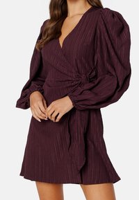 Vestido envolvente burdeos con tejido texturizado, mangas largas abullonadas y un detalle de lazo en la cintura. Corte corto con dobladillo acampanado.