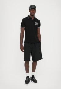 Cămașă polo neagră cu guler alb contrastant și logo, asociată cu pantaloni scurți negri. Modelul poartă adidași negri și o șapcă neagră.