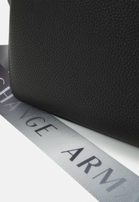 Schwarze, strukturierte Ledertasche mit einer glatten Oberfläche, kombiniert mit einem kontrastierenden grauen Riemen, auf dem der Schriftzug "CHANGE ARM" in fetten schwarzen Buchstaben prangt.