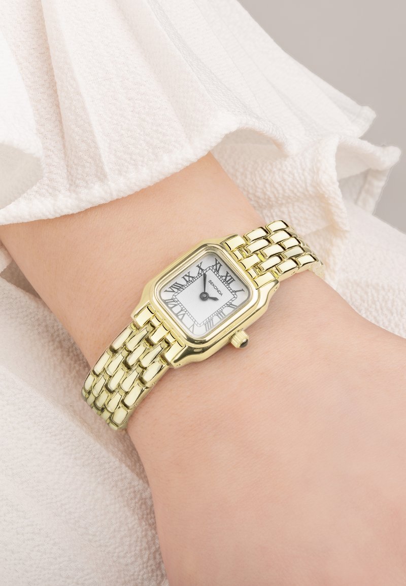 Montre en acier inoxydable doré avec un cadran rectangulaire, un cadran à chiffres romains blancs et un bracelet maillon. Gros plan sur un poignet.